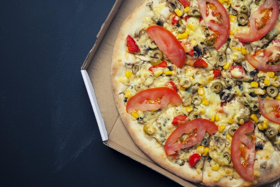 A vegetable pizza with tomato, olives and corn in an open delivery box
